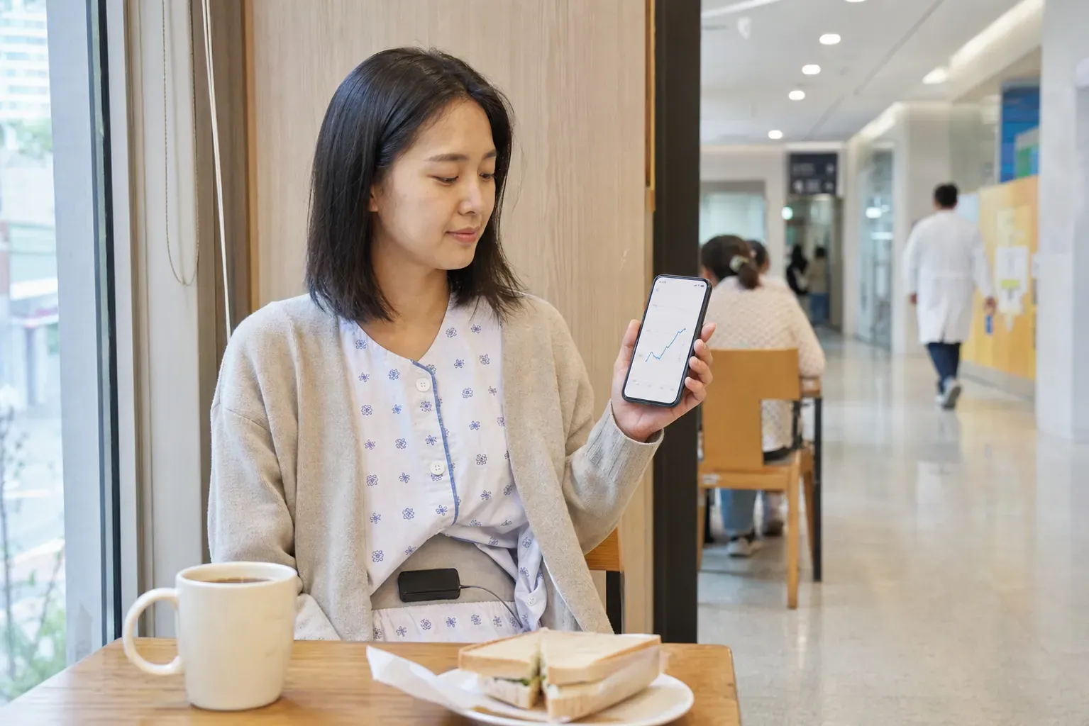 서울아산병원 복도의 작은 카페에서 시범 환자 서지안 씨가 검은색 외부 충전 패치를 옷 위 아랫배에 붙인 채 샌드위치와 커피를 앞에 두고 스마트폰의 완만한 혈당 상승 곡선을 확인하는 모습